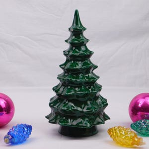 May include: A dark green ceramic Christmas tree with a glossy finish stands among pink and blue ornaments. The tree is surrounded by three pink ornaments and three decorative light bulbs in blue, yellow, and green.