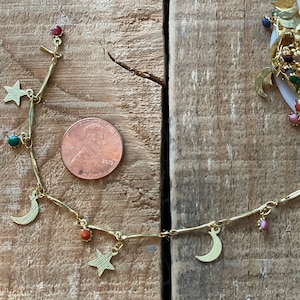 May include: A gold chain necklace with charms of stars, moons, and colorful gemstones. The necklace is displayed on a wooden surface with a US penny for scale.