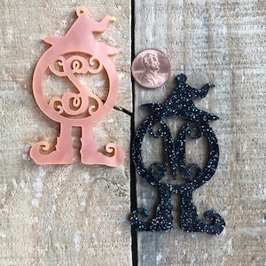 Puede incluir: Dos adornos con temática de Halloween, uno en rosa y otro en purpurina negra. Ambos adornos representan una bruja con un sombrero y un círculo con monograma.