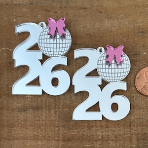 Puede incluir: Dos adornos plateados espejados con el número 26, un detalle de bola de discoteca y lazo rosa. Se incluye una moneda para comparar el tamaño. Los adornos están sobre una superficie de madera marrón.