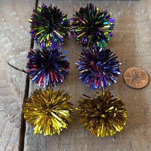 May include: Six colorful pom-poms made of shiny metallic strands. The pom-poms are in three colors: gold, red, white, and blue, and green, purple, and gold.