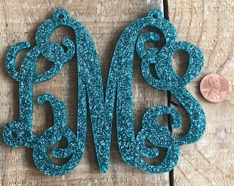 May include: Teal glitter monogram with the letters L, M, and S. The letters are connected in a cursive style and have a small hole at the top for hanging. A penny is next to the monogram for size comparison.