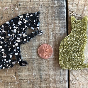 May include: Two glitter cat ornaments, one black with silver glitter and one gold with green glitter. The ornaments are about the size of a US penny.