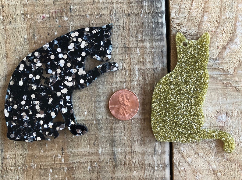 May include: Two glitter cat ornaments, one black with silver glitter and one gold with green glitter. The ornaments are about the size of a US penny.