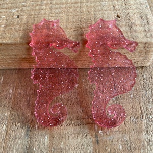 May include: Two pink glitter seahorse earrings. The seahorses are made of a translucent plastic with a glittery finish.