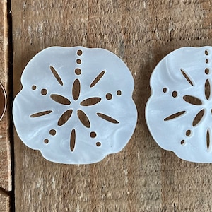 May include: Two white, decorative sand dollar ornaments with a cutout design. The ornaments are about the size of a US penny.