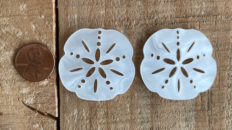 May include: Two white, decorative sand dollar ornaments with a cutout design. The ornaments are about the size of a US penny.