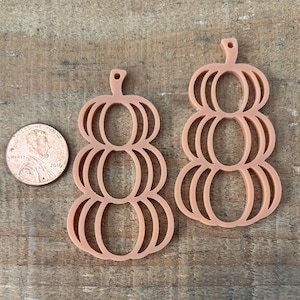 May include: Two pumpkin-shaped cutouts in a light orange color, next to a penny for size comparison. The pumpkins are made of a thin material and have a layered design. The background is a wooden surface.