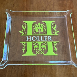 Puede incluir: Una bandeja de vidrio transparente con un diseño de monograma verde neón. El monograma presenta la letra "H" con el nombre "HOLLER" en el centro. La bandeja tiene forma rectangular con bordes curvos.