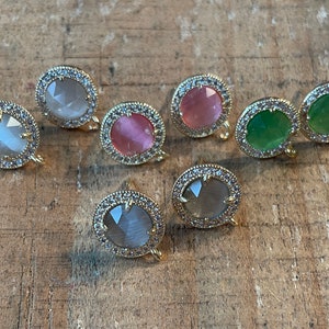 May include: Six pairs of earrings with gold settings and various colored stones. The stones are round and have a sparkly, iridescent finish. The earrings are set in a gold tone metal with a clear crystal halo around the stone.