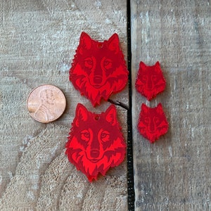 May include: Four red wolf-shaped acrylic charms in varying sizes, with a penny for scale. The charms feature detailed wolf faces and small holes for hanging. The charms are displayed on a wooden surface.