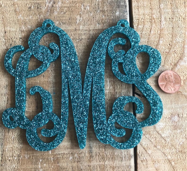 May include: Teal glitter monogram with the letters L, M, and S. The letters are connected in a cursive style and have a small hole at the top for hanging. A penny is next to the monogram for size comparison.