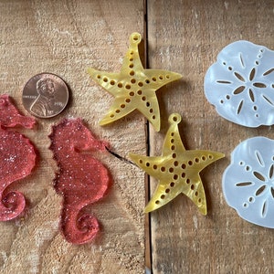 May include: A set of six resin ornaments featuring a beach theme. The set includes two pink glitter seahorses, two yellow starfish, and two white sand dollars.