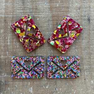 May include: Four glittery acrylic charms in the shape of envelopes and hearts. Two are diamond-shaped with a heart cutout and the other two are rectangular with an X design and heart cutout. The charms are filled with red, pink, yellow, and green glitter.