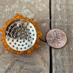 May include: Close-up of the back of a pair of orange beaded earrings. The earrings are about the size of a UK penny.
