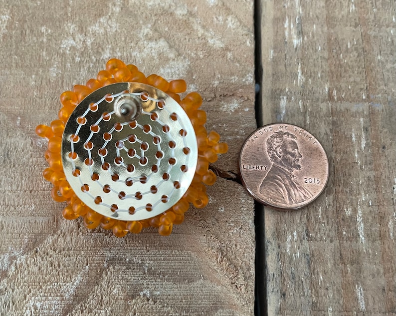 May include: Close-up of the back of a pair of orange beaded earrings. The earrings are about the size of a UK penny.