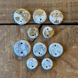 May include: A selection of white ceramic buttons with gold and silver leaf detailing. The round buttons have two holes for sewing. The buttons are of varying sizes and are arranged on a wooden surface.