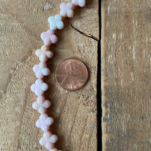 May include: A strand of pink and white shell flower beads on a wooden surface. A US penny is included for scale.