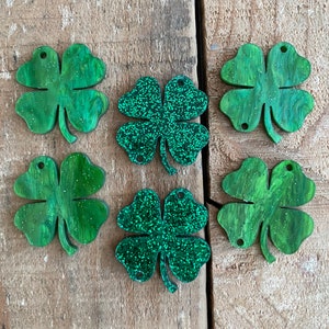 Peut inclure: Six breloques en forme de trèfle à quatre feuilles disposées sur deux rangées. Quatre breloques présentent une finition verte marbrée avec des paillettes, tandis que deux breloques centrales sont recouvertes de paillettes vertes denses.