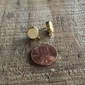 May include: A pair of gold-toned stud earrings with a white sparkly stone. The earrings are shown next to a US penny for size comparison.