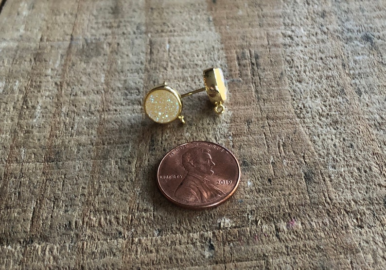 May include: A pair of gold-toned stud earrings with a white sparkly stone. The earrings are shown next to a US penny for size comparison.