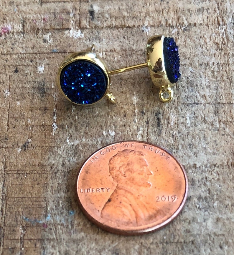 May include: A pair of gold stud earrings with blue glitter round cabochons. The earrings are about the size of a US penny.