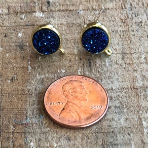 May include: A pair of gold-toned earrings with blue glitter round cabochons. The earrings are about the size of a US penny.