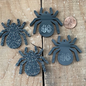 Puede incluir: Cuatro adornos de araña de purpurina negra con un monograma en el centro. Las arañas son aproximadamente del tamaño de una moneda de un centavo de euro.