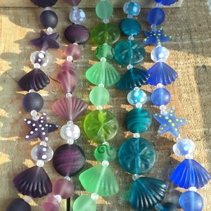 May include: Five strands of glass beads with various sea-themed designs, including seashells, starfish, and sand dollars. The beads are in shades of purple, green, blue, and white.