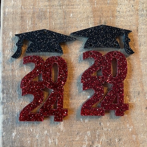May include: Two red glitter graduation year 2024 ornaments with black glitter graduation caps.