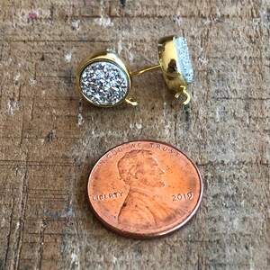 May include: A pair of gold-tone earring posts with a round, clear crystal stone on each post. The stones are covered in tiny, sparkly crystals. The earring posts are about the size of a US penny.