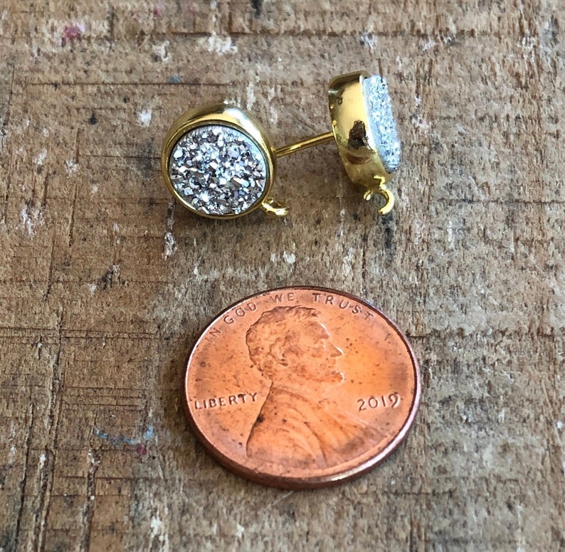 May include: A pair of gold-tone earring posts with a round, clear crystal stone on each post. The stones are covered in tiny, sparkly crystals. The earring posts are about the size of a US penny.