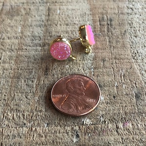 May include: A pair of pink glitter stud earrings with gold posts. The earrings are shown next to a US penny for size comparison.