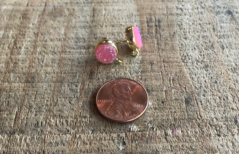 May include: A pair of pink glitter stud earrings with gold posts. The earrings are shown next to a US penny for size comparison.