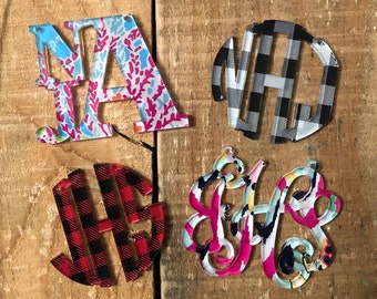 May include: Four acrylic monogram ornaments with different patterns. The first ornament is a light blue and pink floral pattern with the letters "NA". The second ornament is a black and white checkered pattern with the letters "NH". The third ornament is a red and black checkered pattern with the letters "HE". The fourth ornament is a pink, blue, and yellow floral pattern with the letters "LH".