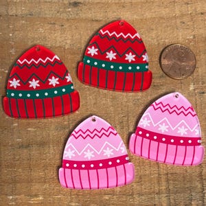 May include: Four acrylic Christmas hat ornaments in red and pink. The red hats have white snowflake and zig-zag patterns, with green and white accents. The pink hats have white snowflake and zig-zag patterns, with red and white accents. A penny is included for size reference.
