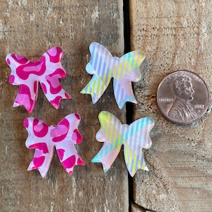 May include: Four clear plastic bows with different patterns. The bows are pink, yellow, and blue with white stripes. The bows are about the size of a US penny.
