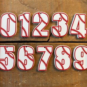 Puede incluir: Diez adornos blancos con temática de béisbol con costuras rojas, cada uno con un número diferente del 0 al 9.