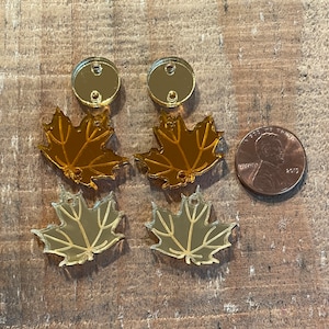 May include: Four gold-colored maple leaf charms with two round gold-colored charms with holes in them. The charms are on a wooden surface.