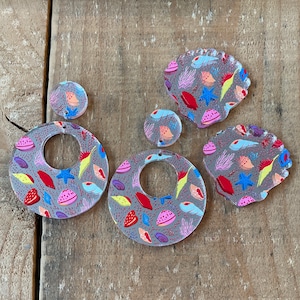 May include: Clear acrylic earrings and pendants with a gray background and a colorful sea life pattern. The designs include round, oval, and seashell shapes, featuring starfish, shells, and coral in red, blue, yellow, and pink.