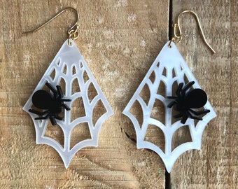 May include: A pair of dangle earrings featuring a white, translucent, diamond-shaped spider web cutout design. Each earring is adorned with a small, solid black spider figure attached to the web. The earrings hang from gold-toned metal hooks.