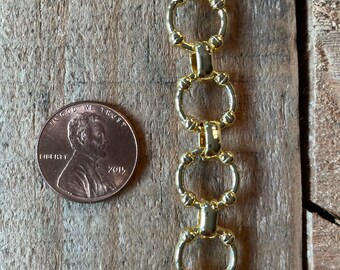 May include: A gold-colored chain with a repeating oval link design. A U.S. penny is next to the chain for scale. The chain is on a weathered wooden surface.