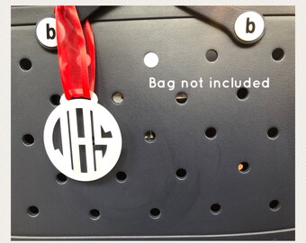 May include: White monogrammed charm with the letters QHS, hanging from a red ribbon. The charm is circular and has a cut-out design. The background is a dark gray surface with several round holes and the text "Bag not included".