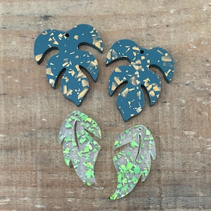 Puede incluir: Cuatro pendientes en forma de hoja. Dos son de color verde azulado con motas doradas, y dos son transparentes con purpurina verde. Los pendientes están colocados sobre una superficie de madera.