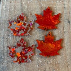 May include: Four maple leaf shaped ornaments, two are clear with orange and red glitter embedded in the resin, and two are solid orange with glitter embedded in the resin.
