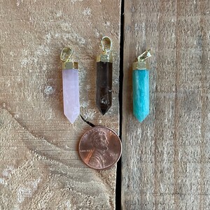 Puede incluir: Tres colgantes de cristal de punta con tapas doradas. Los colgantes son rosa, marrón y azul verdoso.