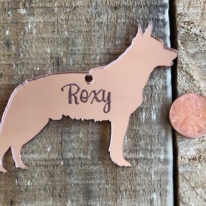 May include: Rose gold metal dog tag with the name "Roxy" engraved on it. The tag is shaped like a German Shepherd and is about the size of a US penny.