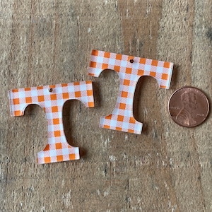 Puede incluir: Dos abalorios de acrílico con forma de letra T en un patrón de cuadros naranja y blanco. Los abalorios son aproximadamente del tamaño de una moneda de un céntimo de euro.