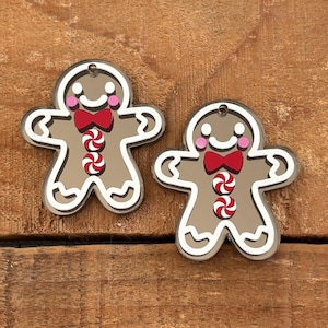 May include: Two gingerbread person ornaments made of brown acrylic with white outlines. Each figure features a red bow tie, two red and white peppermint candy buttons, pink circular cheeks, and a white smile. A small hole is at the top.