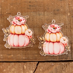 May include: Two clear acrylic charms featuring a stacked pumpkin design. Each charm displays a light orange pumpkin topped with a larger pink pumpkin, accented by pink peony flowers and golden foliage. Each piece has a small hole at the top.